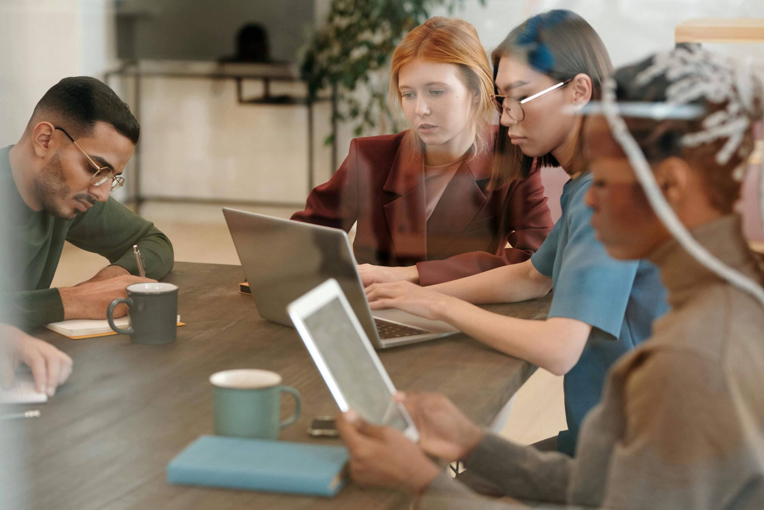 Four people sit at a table working on laptops and tablets in a modern office setting with coffee mugs, collaborating on Medicaid compliance and address verification strategies.