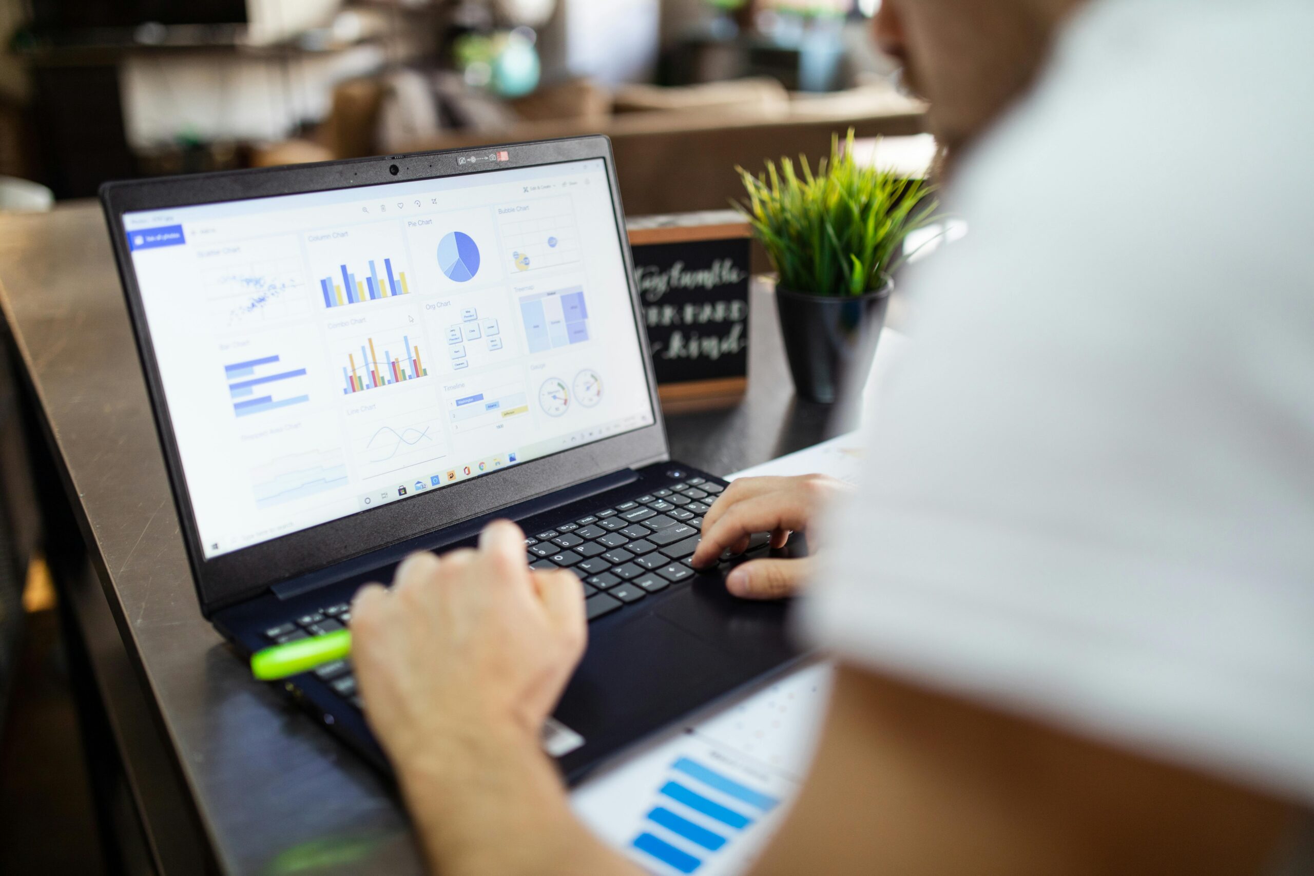 Person analyzing data and charts on a laptop at a desk with papers and a small plant nearby, focused on data tracking to monitor key outcomes in health and housing.