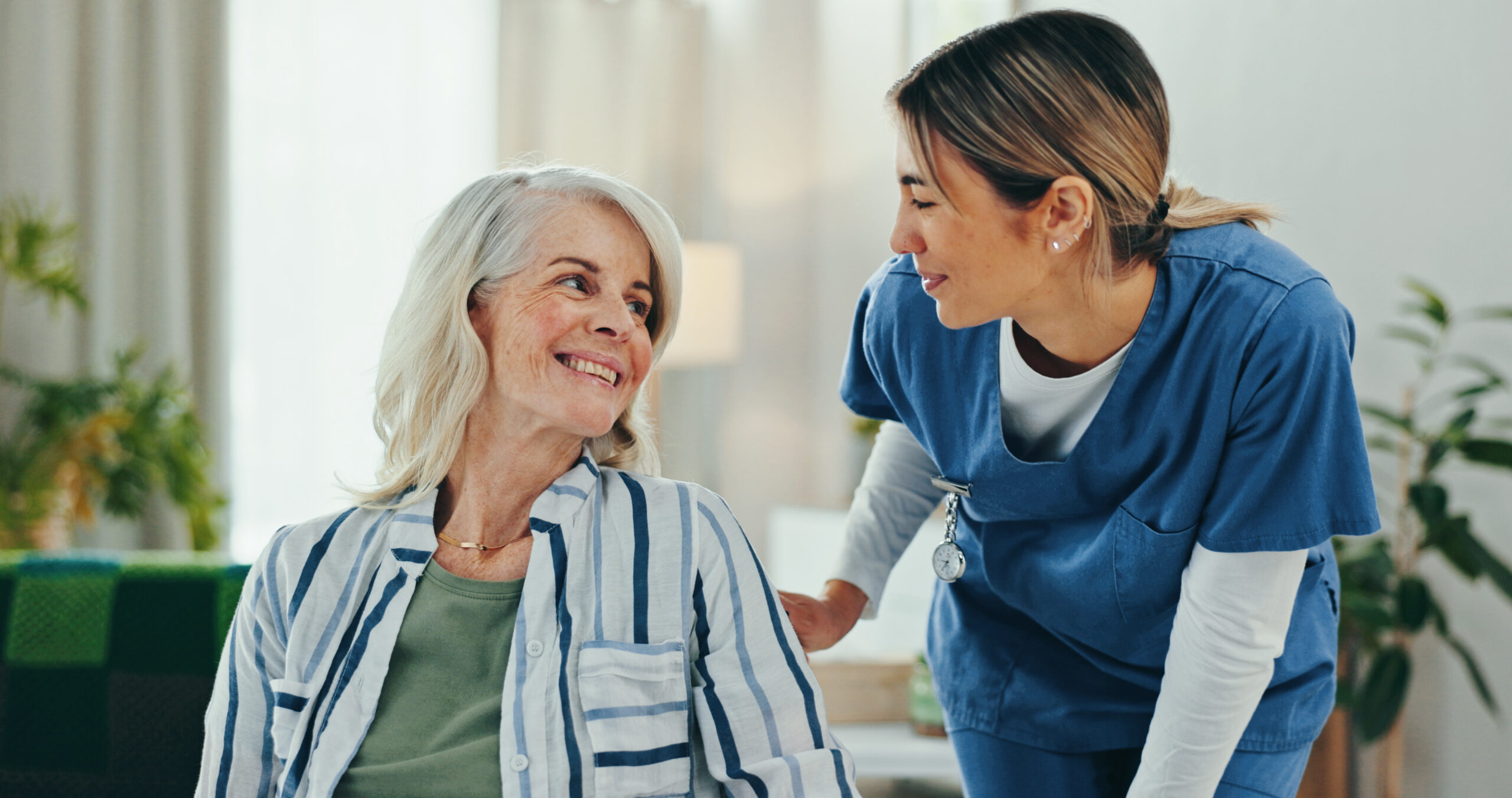 A nurse smiles and talks with an older woman sitting down, both looking happy in a bright room—a heartwarming moment that reflects the impact of Home and Community Based Services.