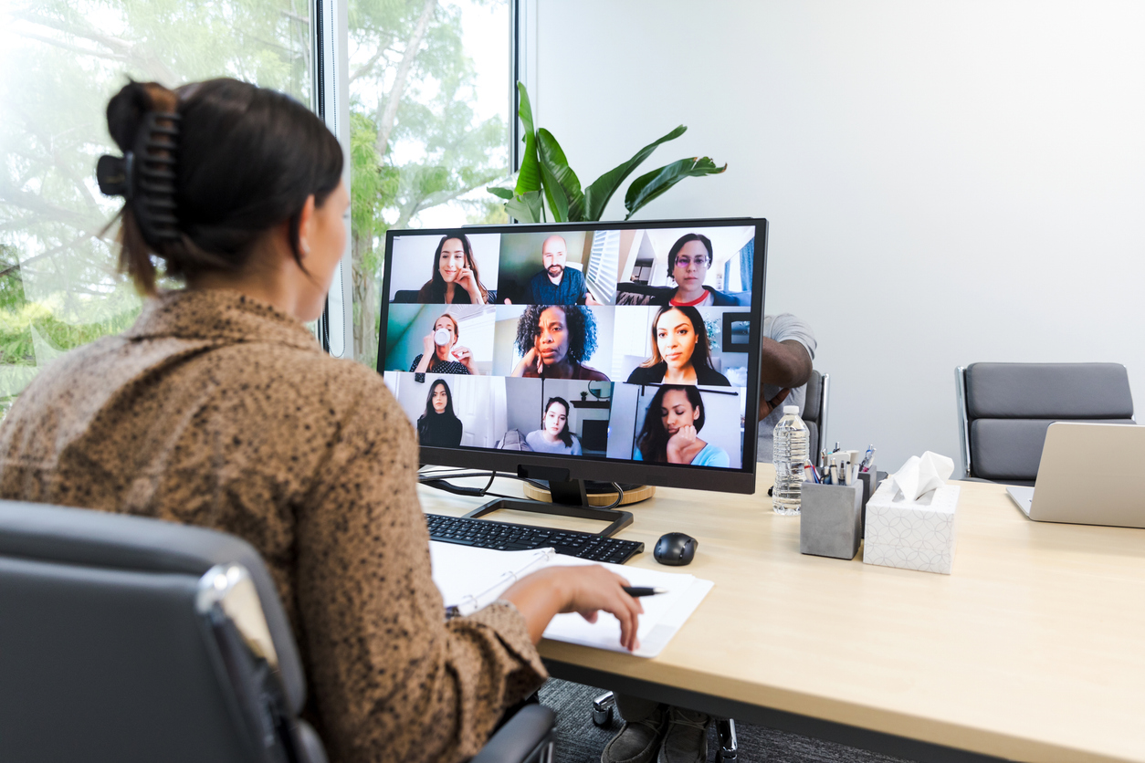 A woman at a desk joins a video conference call on her computer monitor, where ten people discuss topics like H.R.1 Webinar and the Medicaid Program.