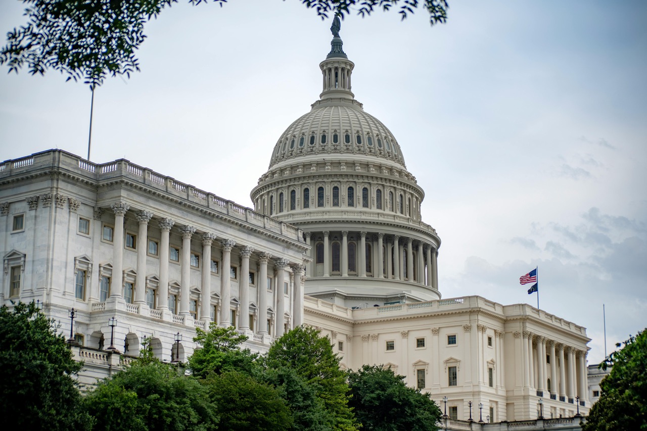 The U.S. Capitol building with its dome and columns stands tall as the American flag flies above, trees in the foreground—a symbol of democracy where key policy solutions are debated and shaped.
