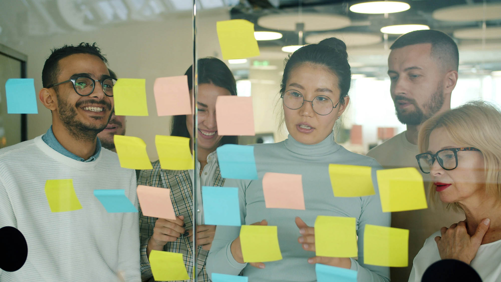 Five people stand in front of a glass wall covered with colorful sticky notes, discussing ideas together on housing and health initiatives at the National Center for Housing & Health in a modern office setting.