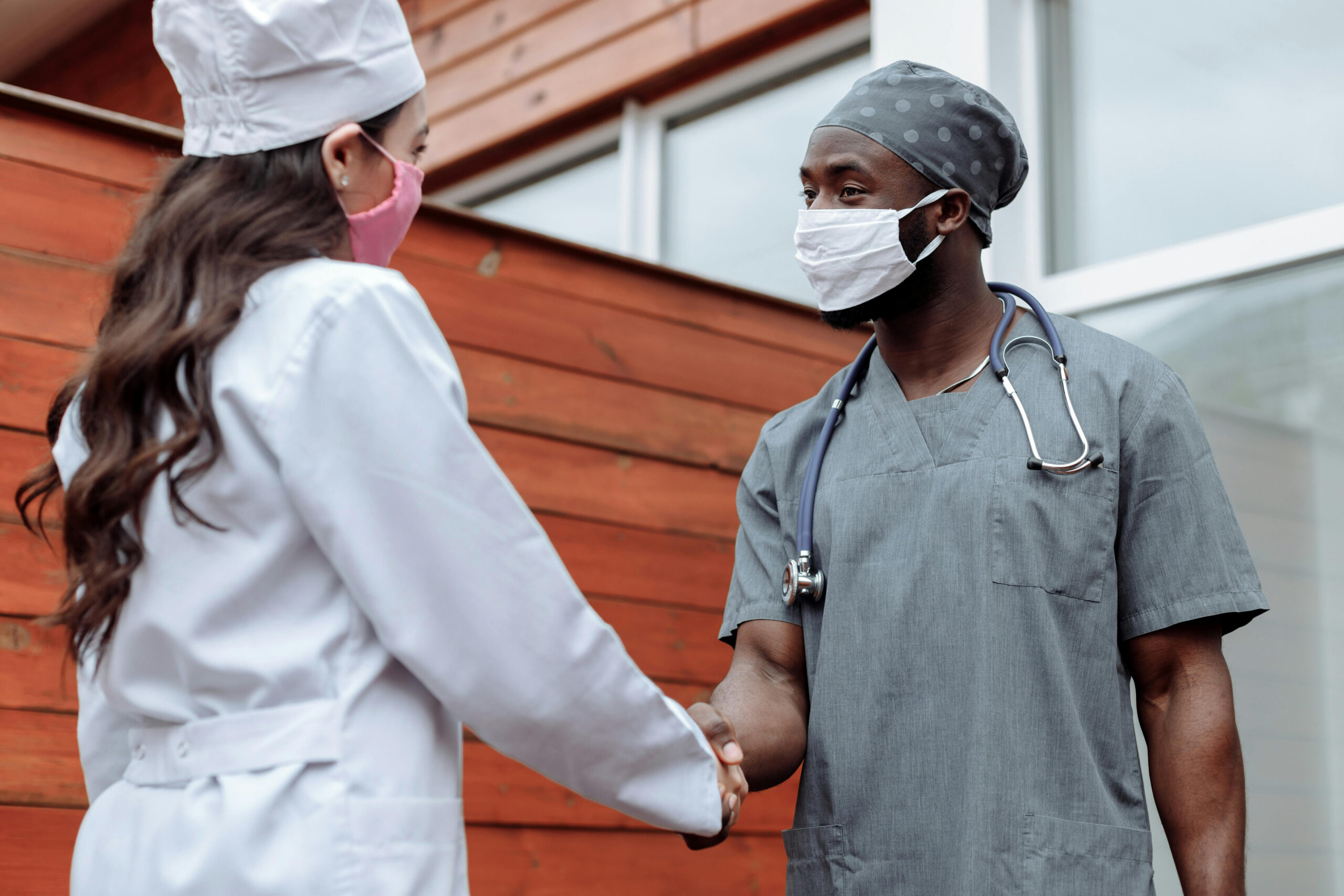Two healthcare workers wearing masks and scrubs shake hands outside a building, standing near crosswalks commonly used by patients covered under Medicaid.