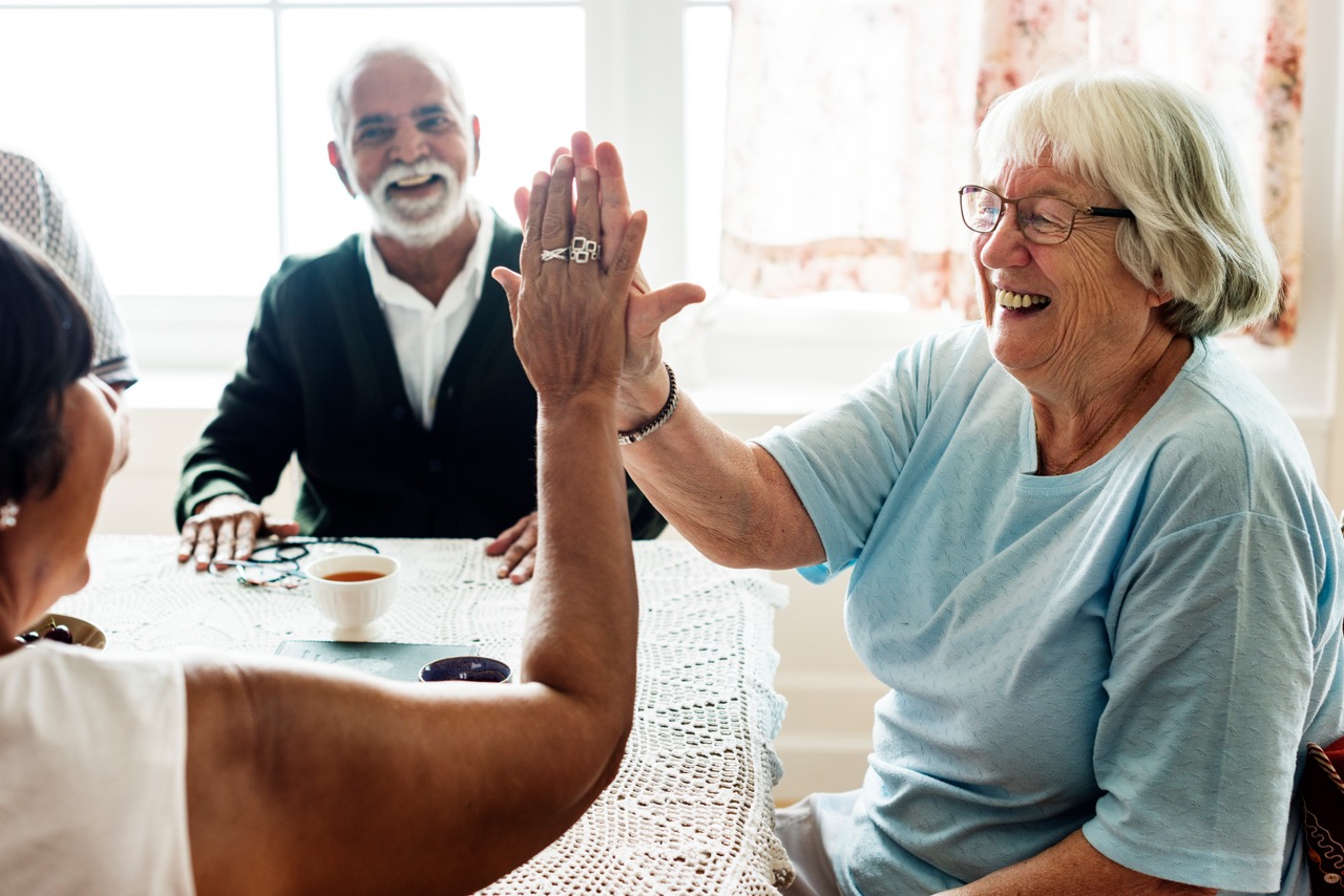 Two elderly women smiling and high-fiving at a table during a training session, with a man smiling in the background.