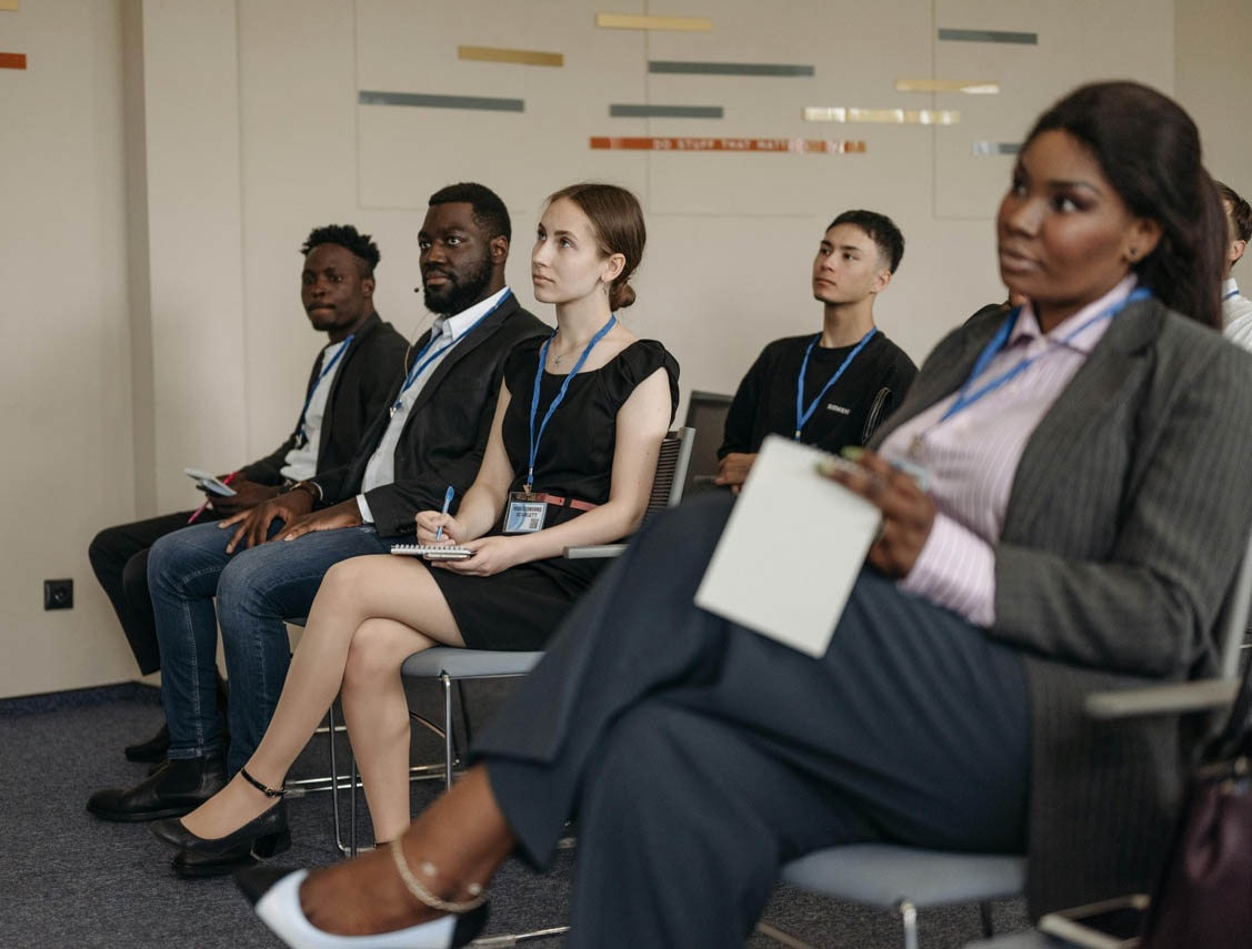 A group of people with notepads sit attentively in a conference room, wearing lanyards, participating in Medicaid training.