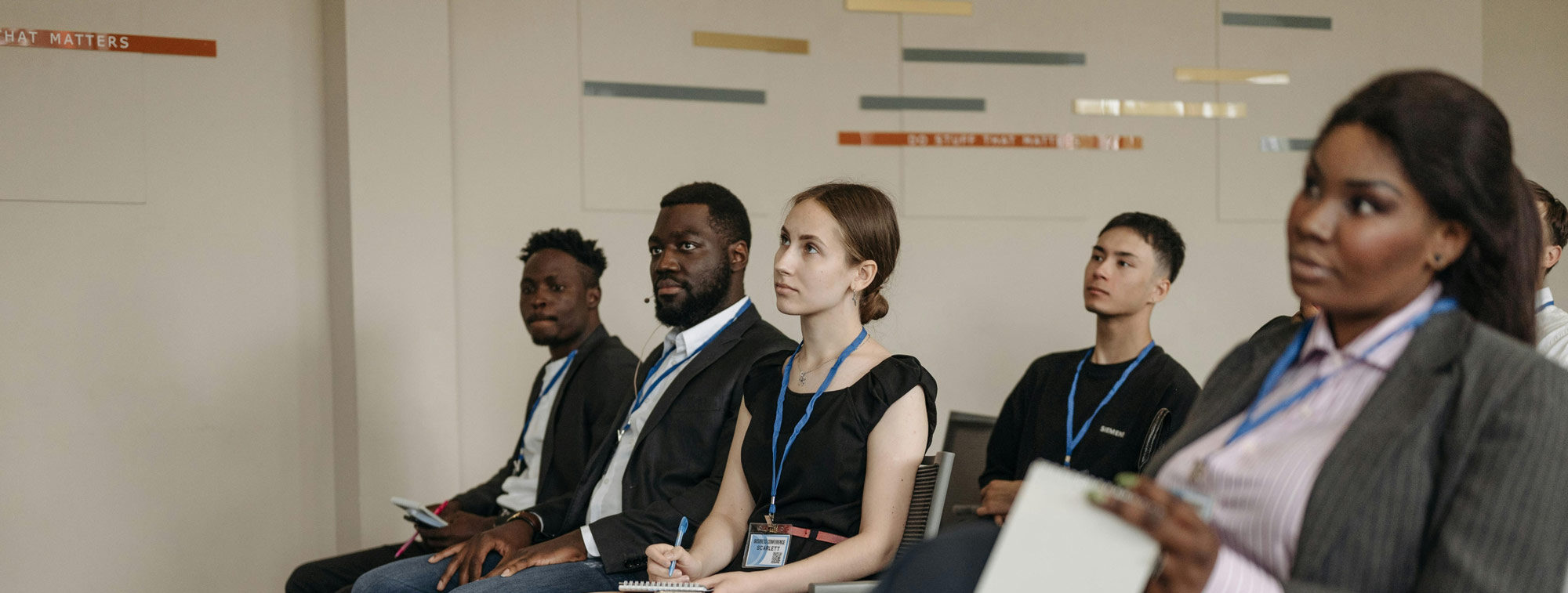 Five people with lanyards sit attentively in a room, listening to a presentation featuring project profiles and case studies.