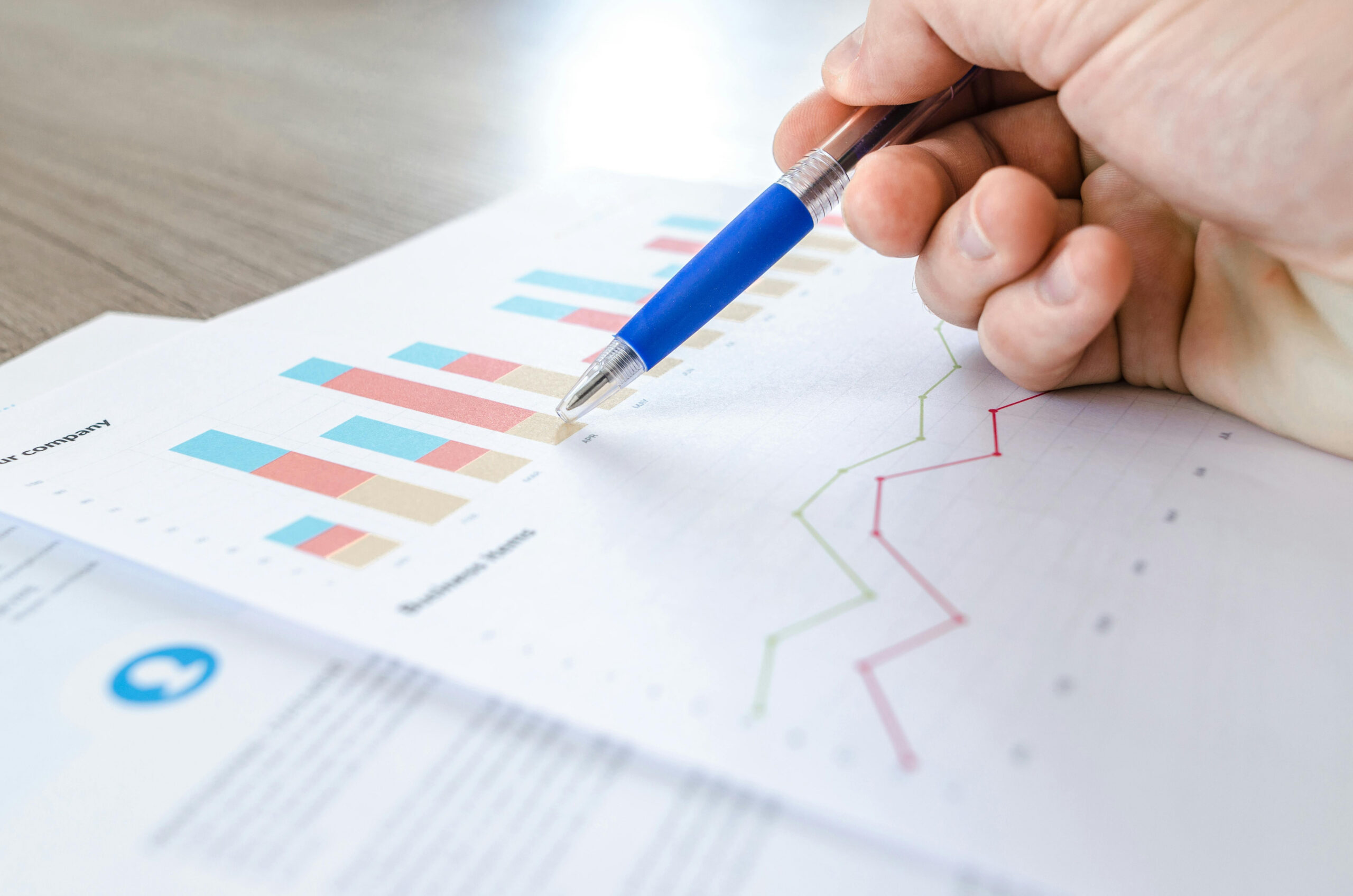 Hand holding a pen pointing at a printed financial chart, highlighting data-driven bar graphs and a line graph on paper to emphasize impact.