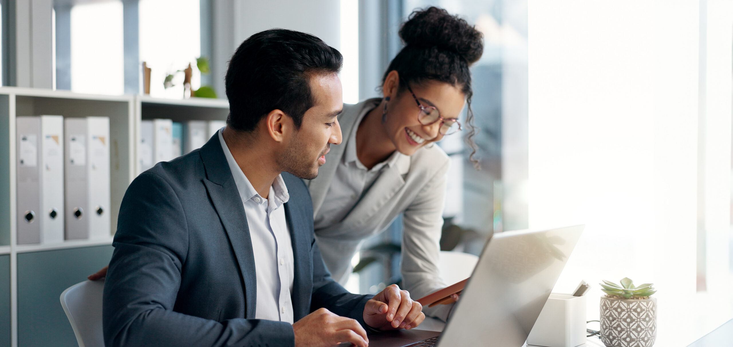 Two colleagues smiling and working together at a desk with a laptop in a bright, modern office.