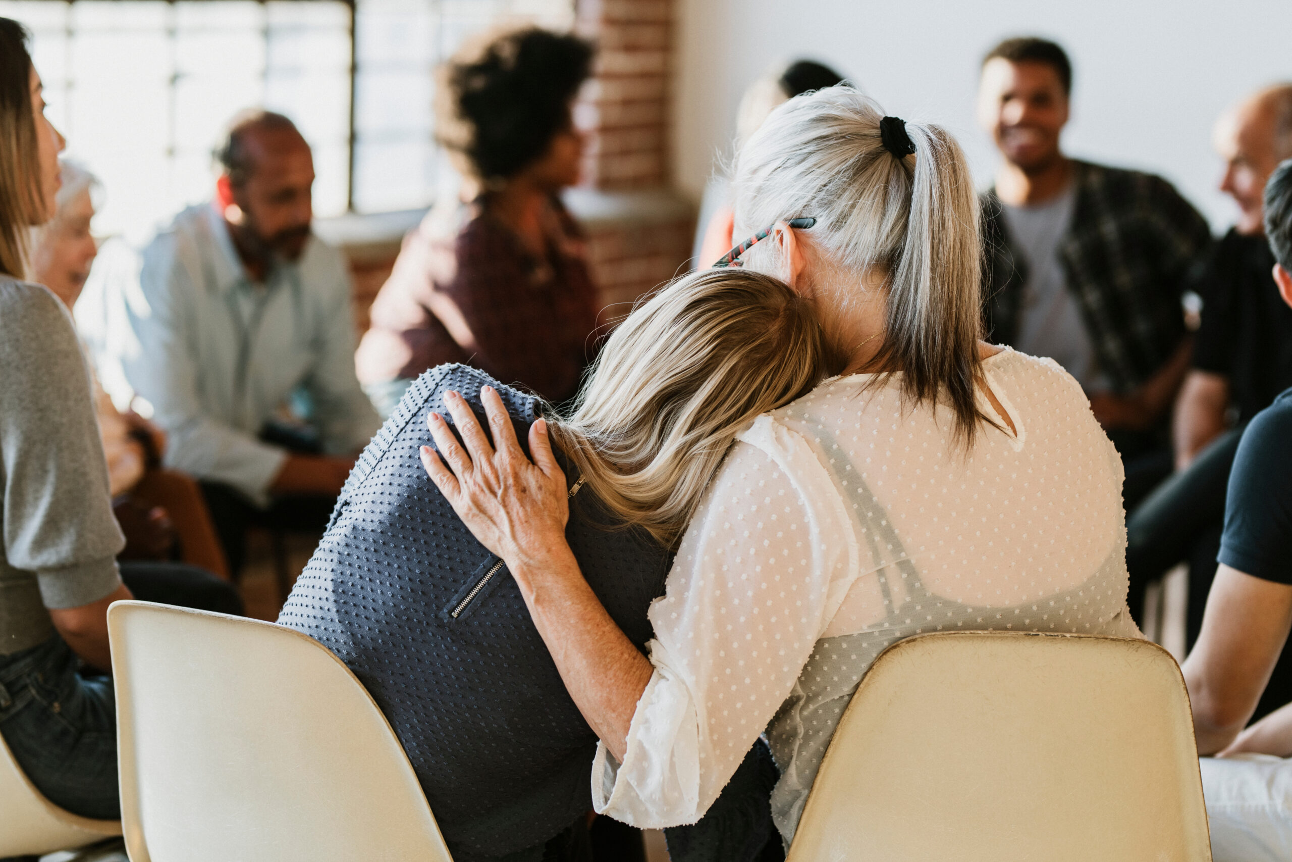 Two women embrace for comfort in a support group, with others seated in a circle in the background.