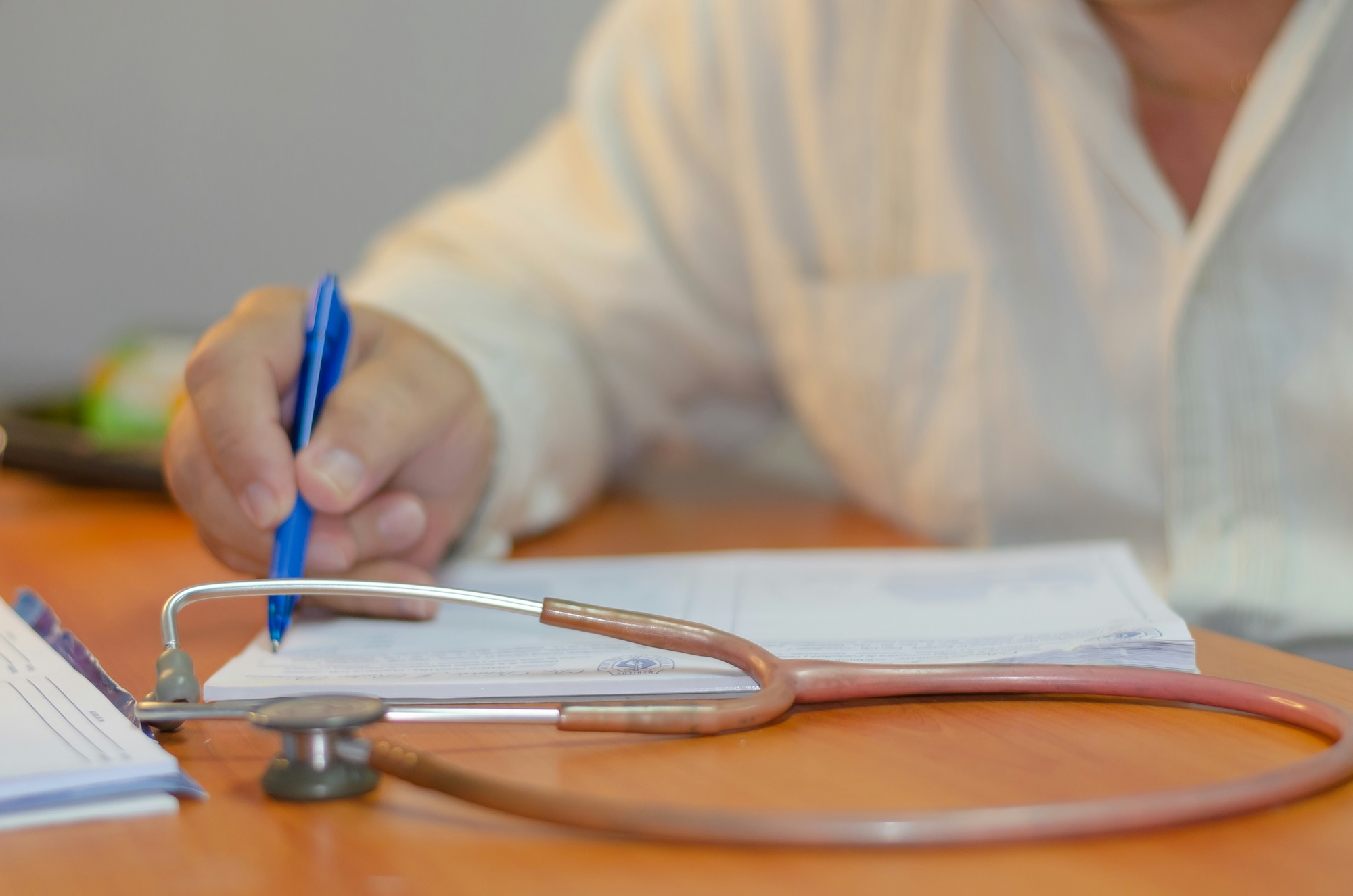 A person writing on paper with a blue pen, a stethoscope lying on the desk in the foreground, possibly filling out Medicaid forms.