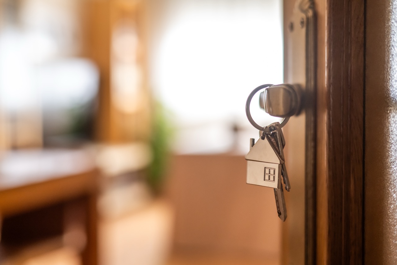 A house-shaped keychain with keys hangs from a door lock in a sunlit, cozy room.