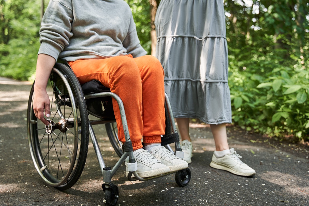 Person in orange pants using a wheelchair outdoors, accompanied by another person standing nearby.