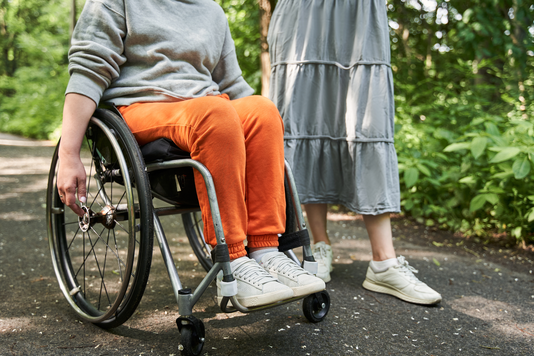 Person in a wheelchair with orange pants beside someone standing on a path in a green park.