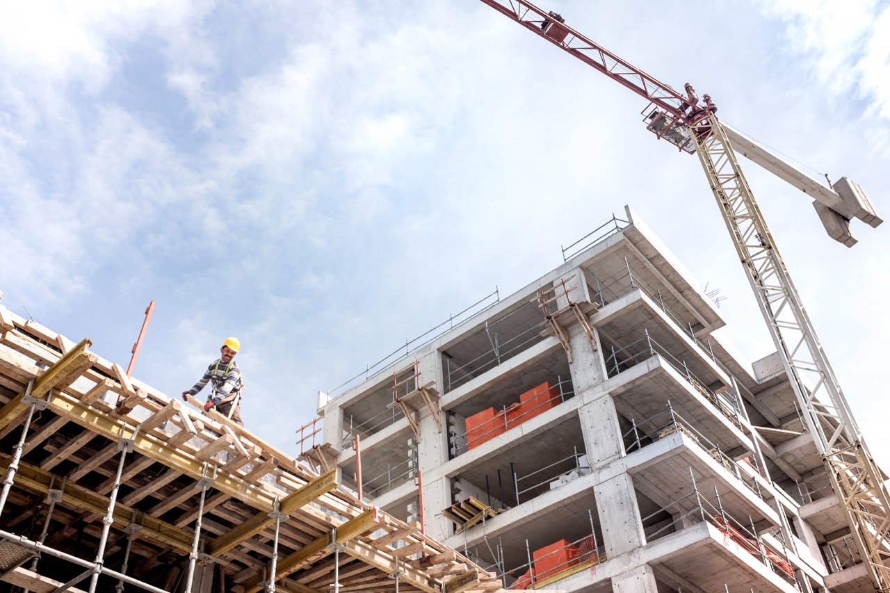Construction worker on scaffolding at a building site with a crane and unfinished concrete structure in the background.