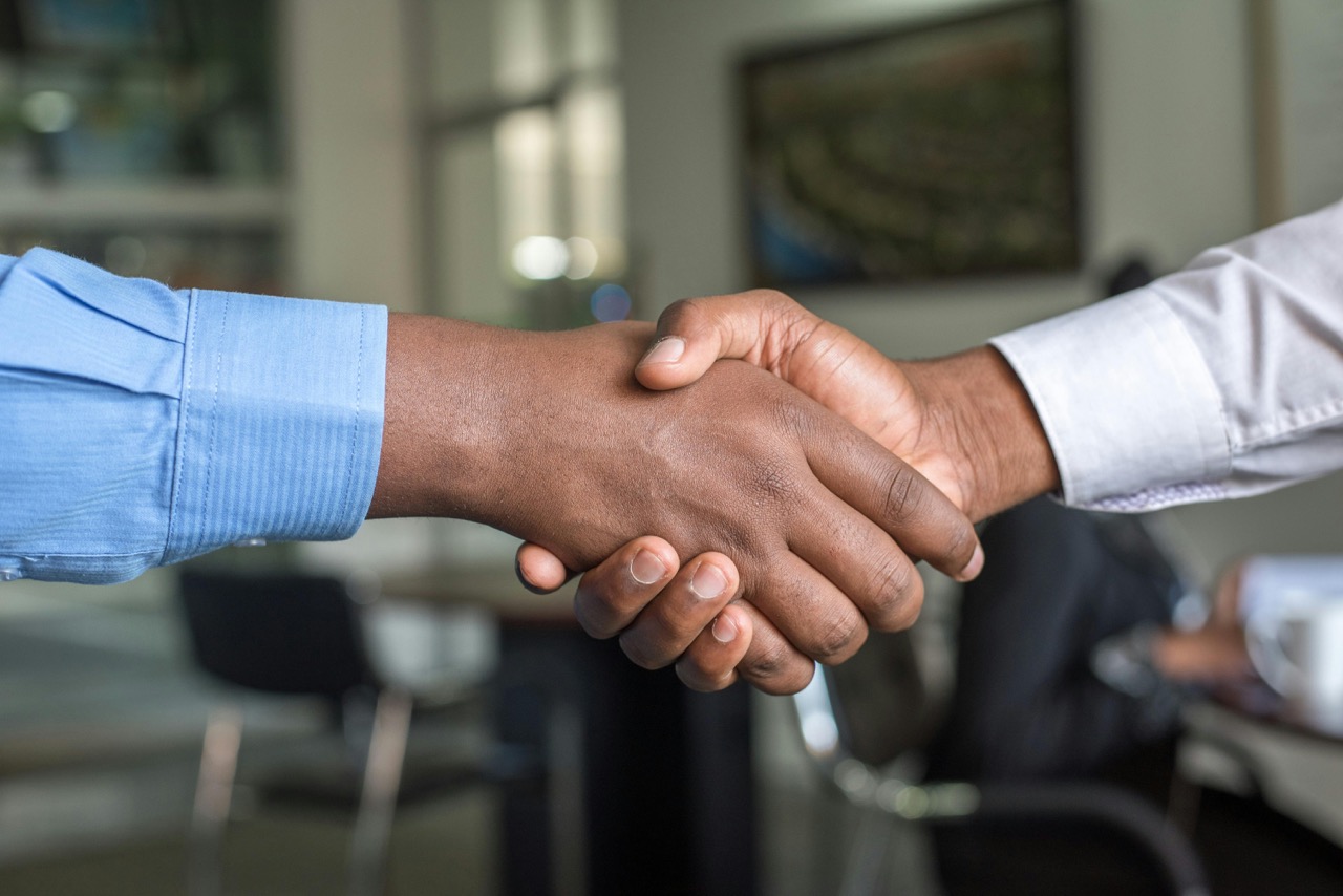 Two people wearing dress shirts shake hands in an office setting, symbolizing agreement or partnership.