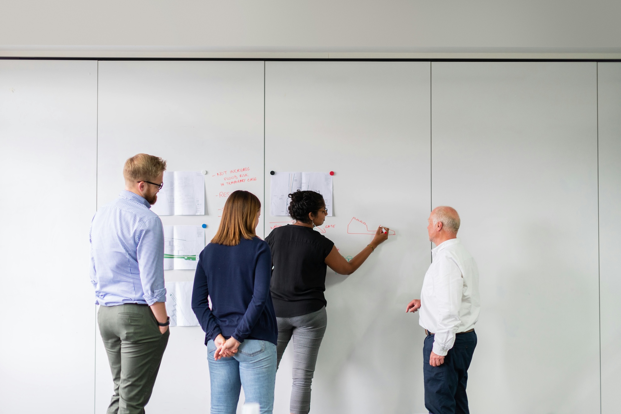Four people stand at a whiteboard; one person is drawing while others watch and discuss.