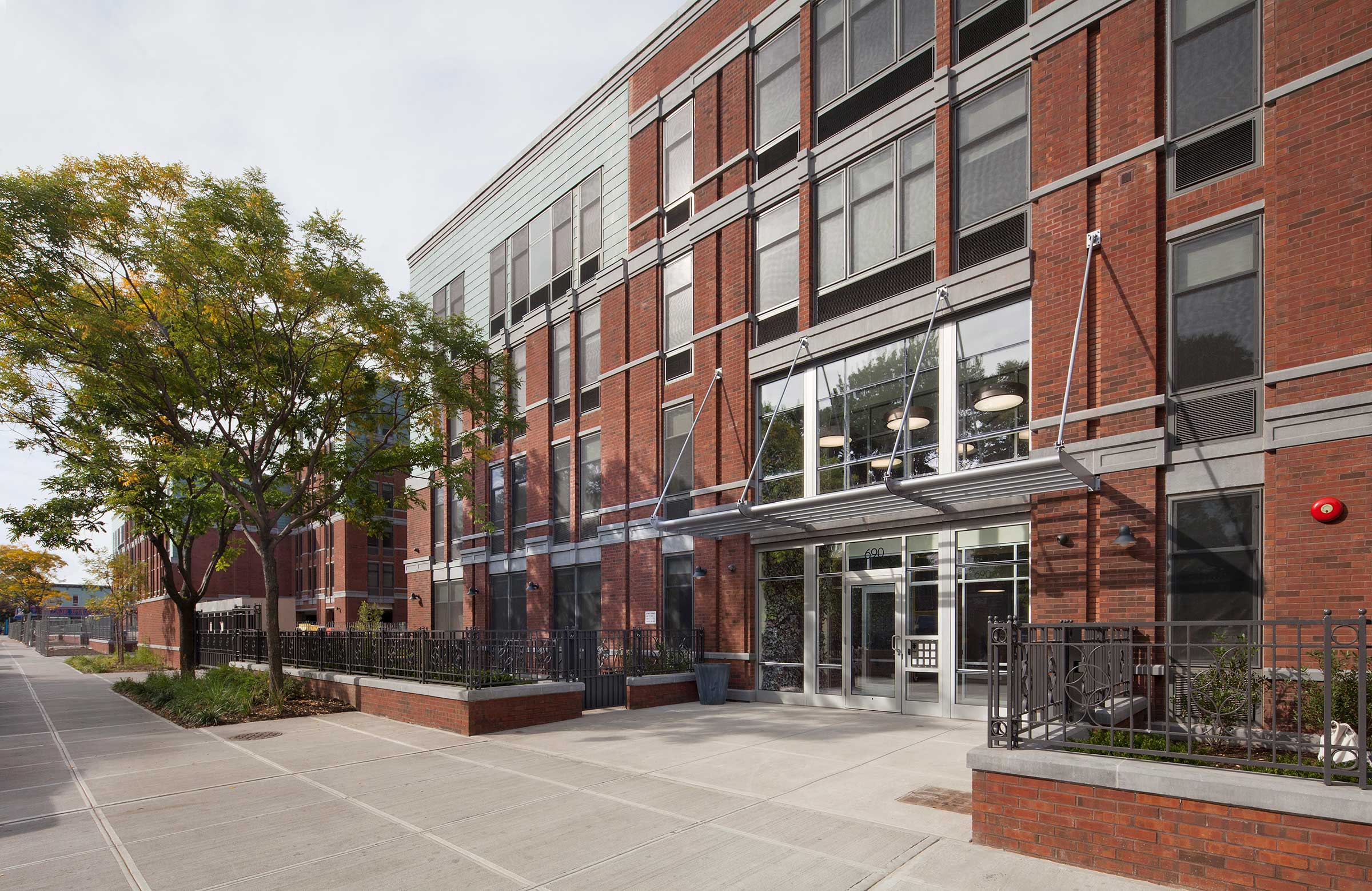 Modern brick building with large windows, tree-lined sidewalk, and metal fencing in an urban setting, part of the CAMBA Gardens II development.