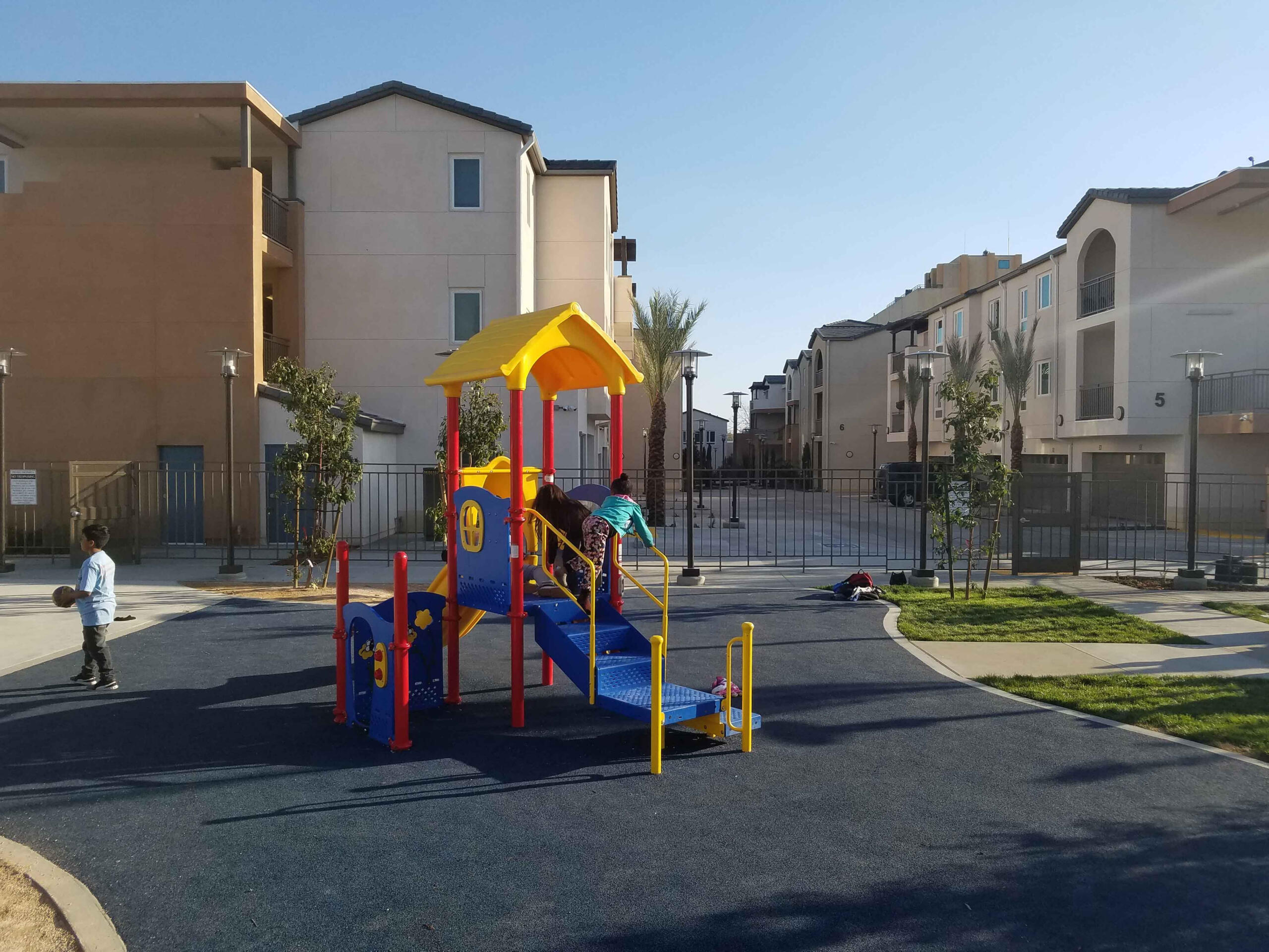 Children playing on a colorful playground structure at Holt Family Apartments in a residential neighborhood on a sunny day.