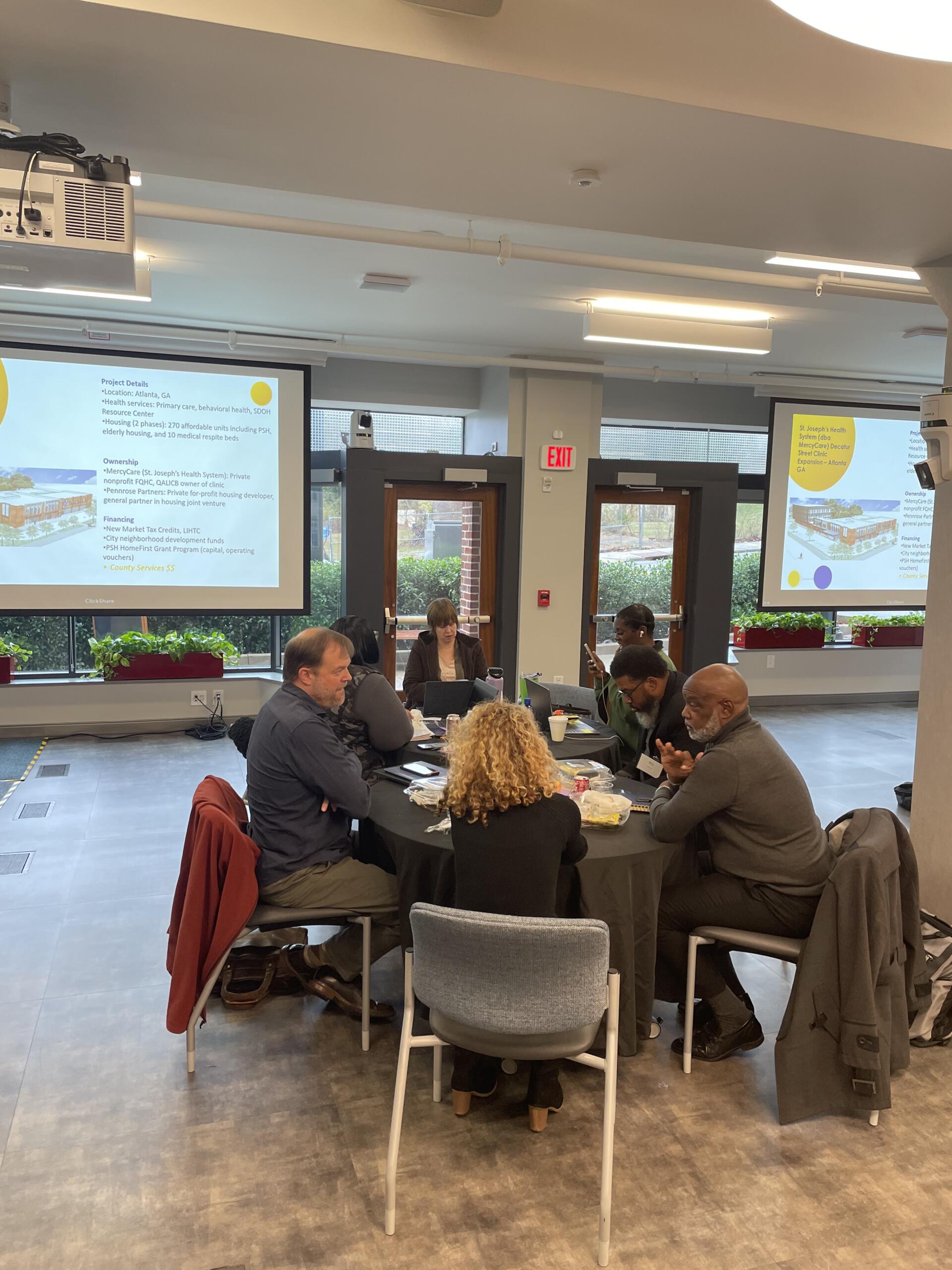 Six people sit around a table in a conference room with presentation slides projected on screens.