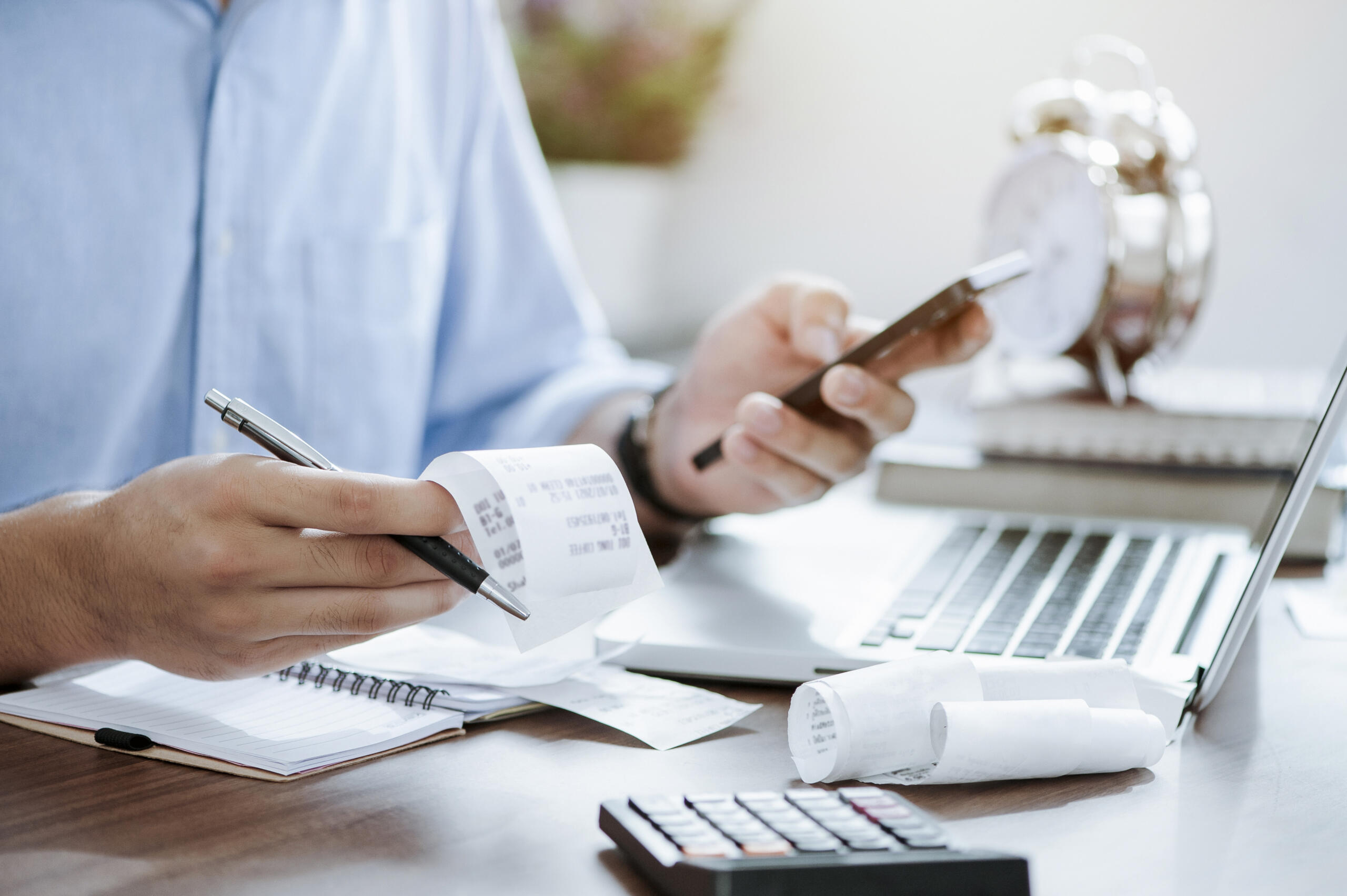 Person holding receipts and a phone, working at a desk with a laptop, calculator, and notepad—managing finances for supportive housing or tracking healthcare investments, possibly preparing documents for New Markets Tax Credit applications.