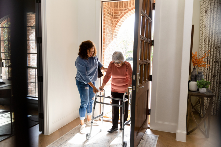 A woman helps an elderly woman using a walker step inside a bright, sunlit home.