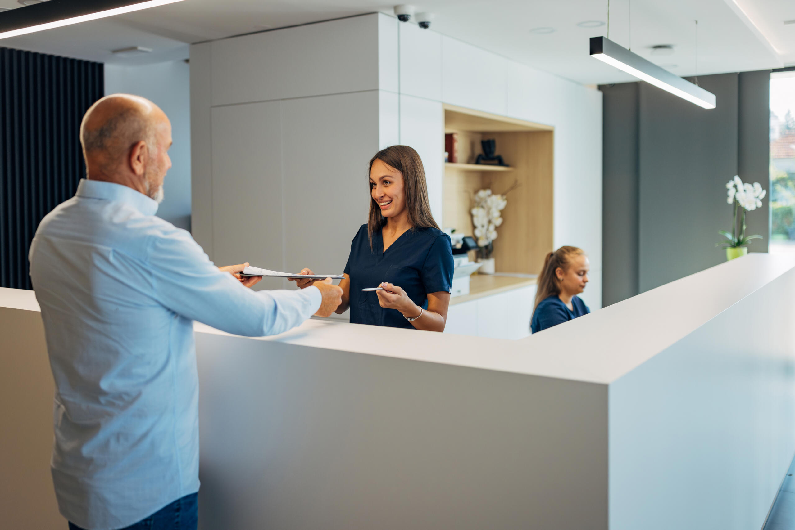 A smiling receptionist hands paperwork to a man at a modern clinic front desk, with another staff member in the background, highlighting the welcoming atmosphere of this National Center for health services.