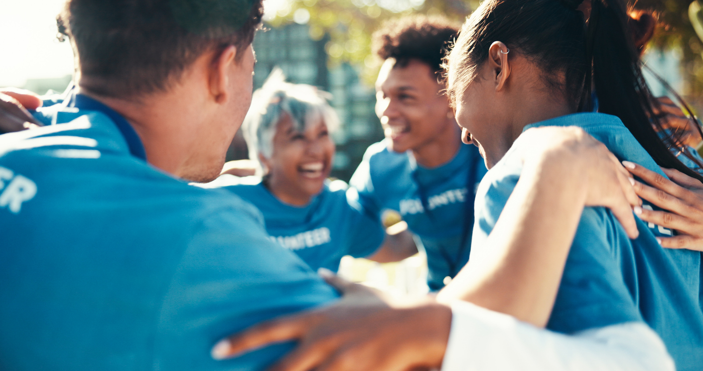 Four volunteers in blue shirts smiling and hugging in a cheerful group huddle outdoors.