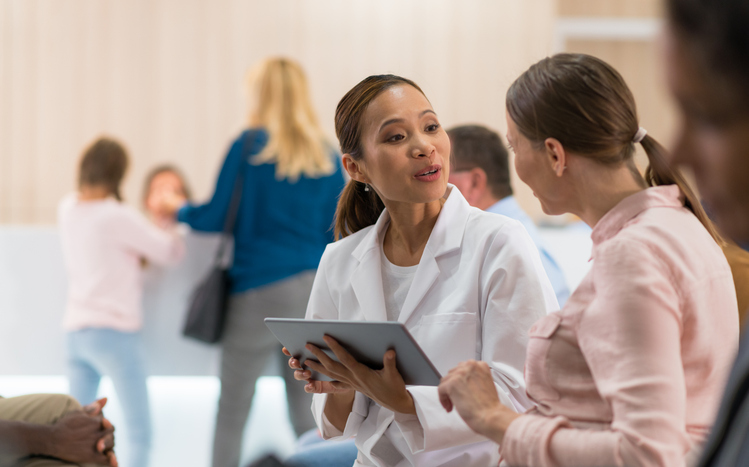 A female doctor with a tablet talks to a patient in a waiting room with people in the background.